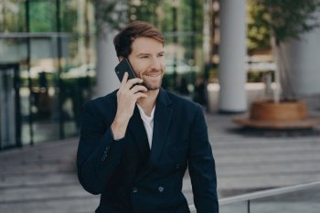 Confident entrepreneur in formal attire makes phone call, enjoying conversation during work break near office building.