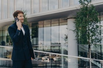 Bearded man in black suit makes successful project call on smartphone near office building, combining job and technology.