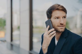 Pensive male entrepreneur in black jacket makes call, holds mobile interview, communicates with customer or partner, focused and professional.