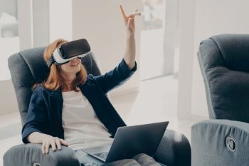 Impressed redhead businesswoman using VR glasses for remote work, interacting with 3D objects and pointing in the air while working on her laptop from home.