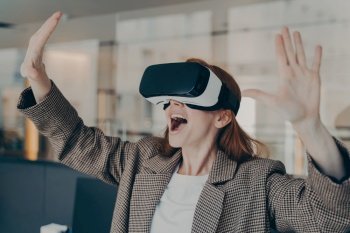 Excited businesswoman in VR glasses, amazed by virtual reality experience in office, expressive gestures and joyful expression.