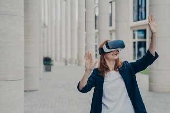Ginger businesswoman in formal attire using VR headset outdoors, exploring virtual world for business purposes.