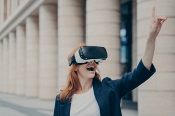 Redhead businesswoman amazed by VR experience outdoors, smiling and attempting to interact with virtual objects, blurred buildings in the background.