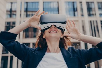 Immersed in virtual reality, futuristic redhead businesswoman wearing 3D goggles stands outside, looking up with awe, holding the portable VR glasses with both hands.