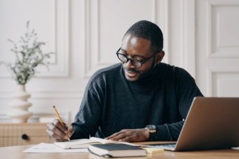 Confident Afro-American businessman working remotely, preparing monthly financial report at home office, taking notes and analyzing charts on laptop.