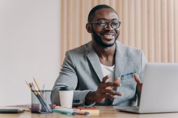 Joyful Afro American businessman in glasses and a stylish blazer leading a remote meeting, participating in an online conference with a bright smile on his face.