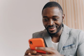 Smiling African self-employed man in suit, chatting with partners on smartphone, sending emails before business meeting.