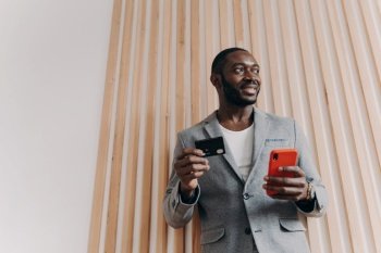 Excited African-American businessman shopping online, using smartphone and credit card, ordering food and making a long-awaited purchase via e-banking.