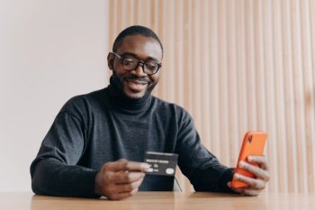 Cheerful African-American entrepreneur making online orders from home office, using smartphone and credit card for seamless e-commerce transactions.