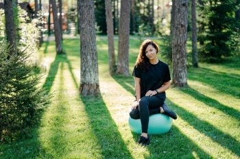 Active brunette woman resting after exercise, urban environment, green grass, trees, sunny, practicing gymnastics, leading active lifestyle.