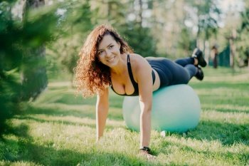 Happy sportswoman in active wear uses gymnastic ball for outdoor fitness, posing on green grass with a smile.