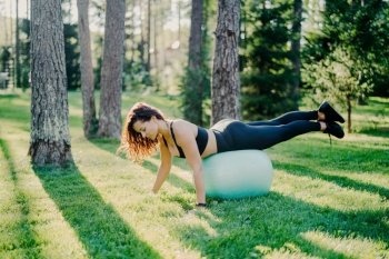 Sporty woman does gymnastic exercises outdoors, leaning on fitness ball, wearing cropped top, leggings, sneakers, working out in forest, poses on green grass near trees.