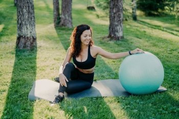 Active woman takes break after exercises, poses on fitball with fitness mat in forest. Perfect body shape, fitness equipment.
