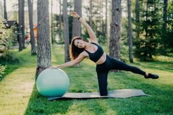 Active woman in sportswear does exercises with fitness ball, posing outdoors against tall trees on a mat.