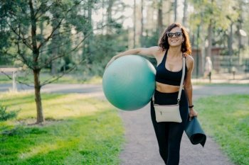 Happy brunette sportswoman in active wear and sunglasses walks outdoors, enjoying fresh air and nature, carrying fitness ball and mat.