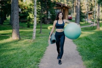 Sportswoman carries fitness ball and mat, walks on road amidst trees and grass, embracing healthy lifestyle.