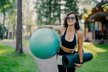 Joyful female pilates instructor, holding fitness ball and rolled-up mat, smiles outdoors in beautiful nature, enjoying a sunny summer day.