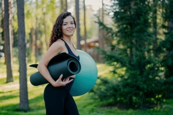 Curly brunette carries fitness gear, poses in forest or park, has outdoor fitness training, promotes active lifestyle.