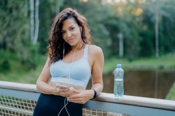 Athletic brunette in top and leggings on bridge, listening to music, using phone, staying hydrated. Active lifestyle.
