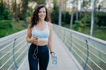 Active woman in sportswear with phone and earphones, taking break after jogging, holding water bottle at bridge. Fitness lifestyle.