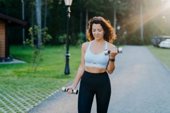 Motivated brunette woman in sports bra and leggings trains biceps with dumbbells outdoors. Fitness exercise concept.
