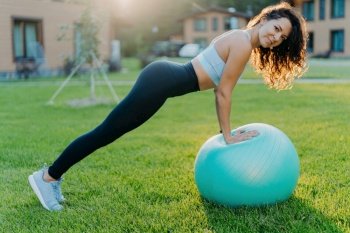 Slim woman exercises with fitness ball, wears sportswear, poses on green lawn, enjoys outdoor training. Pilates and yoga.