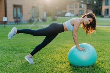 Brunette woman on fitness ball does gymnastic exercises, wears sportswear, poses outdoors, breathes fresh air.