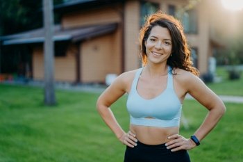 Beautiful curly-haired woman, in good shape, smiles with hands on waist, poses outdoor against house. Morning workout.