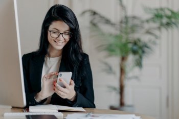 Smiling female freelancer works remotely, focused on smartphone, dressed formally, in a coworking space.