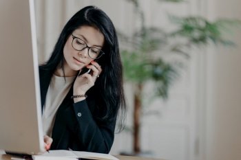 Stylish female entrepreneur in black suit, confidently talking on the phone, taking notes, posing at the desk in an office.