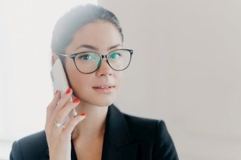 Close-up of confident brunette woman in black suit, talking on mobile with a serious expression, wearing glasses.