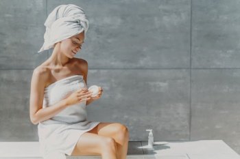 Relaxed woman applies lotion after shower, poses in bathroom, wrapped in towel, enjoys spa treatments.
