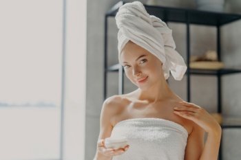 Satisfied woman applies cream, healthy skin, towel wrapped, smiles.