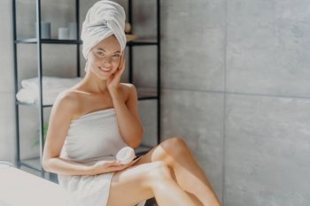 Smiling woman applies facial cream, wrapped in towel, bathroom.