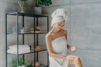 Soft skin after bath. Woman applies cream, wrapped in towel. Beauty and wellness in bathroom.