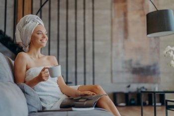 Relaxed woman enjoys tea, reads magazine on sofa. Well cared, healthy skin. Casual and leisure concept.