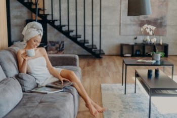 Relaxed woman enjoys tea, wrapped in towel on sofa. Cozy home interior, beauty, relaxation concept.
