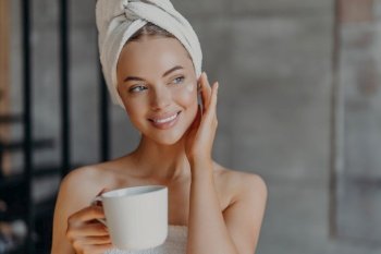 Pretty woman applies face cream, minimal makeup, wrapped in bath towel, enjoys coffee at home. Beauty, relaxation concept.