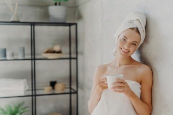 Smiling woman wrapped in towel, drinks coffee, enjoys indoor spa. Wellness, relaxation concept.