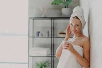 Pleased woman enjoys spa with glowing skin, towel-wrapped, drinks tea in cozy bathroom. Beauty and relaxation.