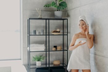 Beautiful woman applies face mask, wrapped in towel, in cozy bathroom. Skin care concept.