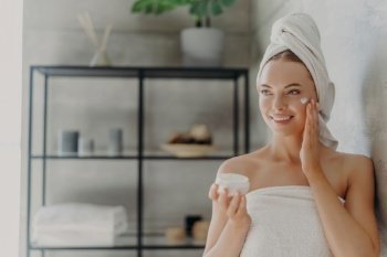 Photo: Pretty woman applies face cream, wrapped in towel, stands in bathroom. Skin care concept.