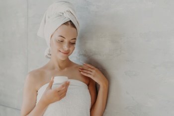 Satisfied woman applies face cream, wrapped in towel, near grey background. Sauna morning routine.