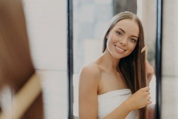 Beautiful woman smiles and combs her straight hair, wrapped in towel, in bathroom. Women’s beauty concept