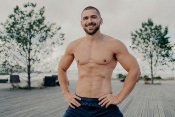 Happy muscular man with bristle poses confidently, hands on waist, enjoys outdoor setting. Fitness, positive mindset.