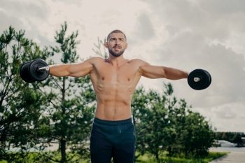 Muscular bodybuilder trains outdoors, lifts weights. Biceps workout in nature.