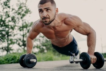 Muscular man does push-ups, focuses outdoors, trains abs with barbells. Sporty lifestyle.
