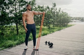 Strong bearded athlete uses resistance band for outdoor workout, performs exercises with sport equipment.