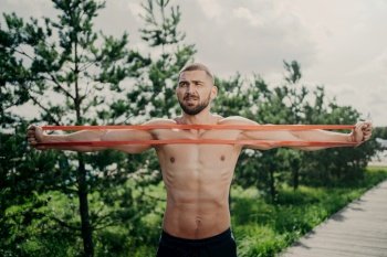 Sporty man with bare chest stretches elastic expander, builds strong muscles, poses outdoors.