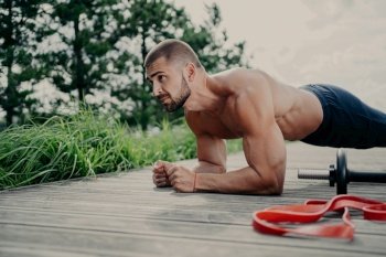 Fit bearded man does plank exercise outdoors, shows endurance and motivation, poses near sports equipment.